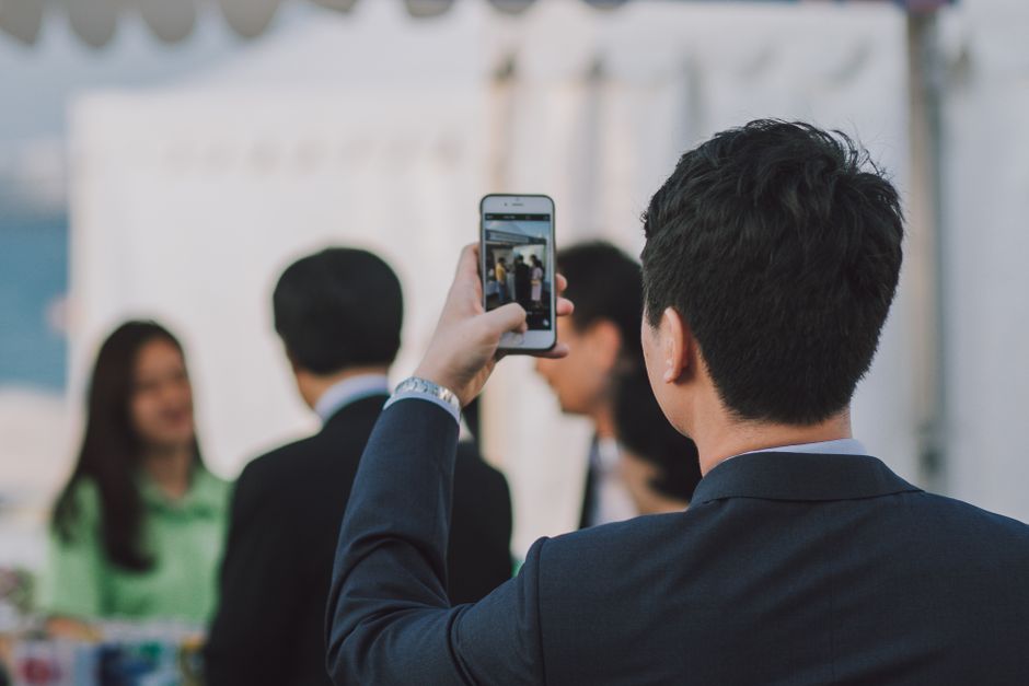 Homem de terno fotografando colegas com smartphone durante evento de negócios ao ar livre.