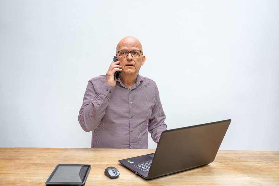 Empresário sênior usando smartphone e laptop na mesa do escritório, engajado em uma chamada.